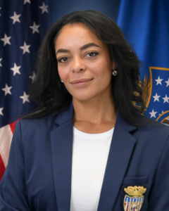 A woman with long dark hair in a blue blazer and white shirt poses in front of American flags, smiling slightly.