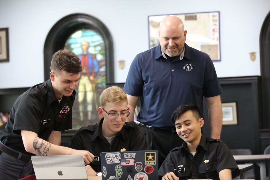 Four cadets in uniform sit around a table looking at a laptop with stickers, while an adult man stands behind them smiling. A stained glass window and framed items are visible in the background at Valley Forge Military College, highlighting leadership.