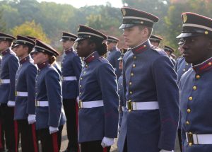 A group of cadets in blue uniforms and hats stand in formation outdoors at Valley Forge Military College, demonstrating leadership during a ceremonial event. Trees and greenery are visible in the background.