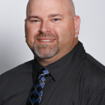 A bald man with a goatee smiles at the camera. He is wearing a black collared shirt and a blue and black patterned tie, reflecting the leadership qualities instilled at Valley Forge Military College, set against a plain light background.