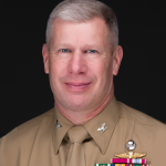 A middle-aged man in a tan military uniform with short gray hair, smiling, displays colorful medals and badges—exuding the leadership of a distinguished Valley Forge Military College cadet—against a black background.