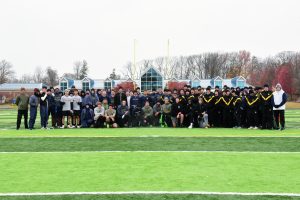 A large group of cadets in athletic wear pose together on an outdoor sports field with green turf. Trees and a blue building with triangular windows highlight the Valley Forge Military College campus in the background.