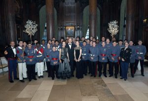 A large group of cadets and young people in formal military-style uniforms and evening gowns stand posed indoors in an ornate room at Valley Forge Military College, with wood paneling, columns, and decorative trees, looking toward the camera.