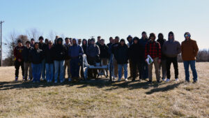 A group of about 30 people, including Cadets, stand outside on a grassy field gathered around a historical cannon. Most are wearing jackets and hoodies under the clear blue sky, reflecting the spirit of leadership at Valley Forge Military College.