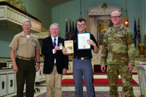 Four men stand smiling indoors, two in military uniforms and two in suits. The cadet second from right holds an award certificate and a book, showcasing leadership at Valley Forge Military College, while the man second from left holds a small medal-like object.