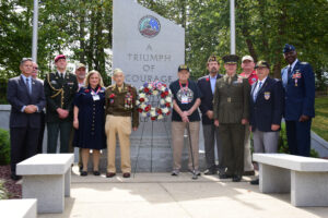 A group of veterans, officials, and a Valley Forge Military College cadet stand in front of a memorial stone engraved with A Triumph of Courage. Wearing uniforms and medals, they pose by the wreath centered at the memorial, honoring leadership and service.