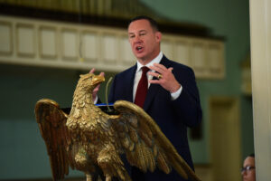 A man in a dark suit and red tie speaks passionately at a podium shaped like a large golden eagle, embodying the leadership values of Valley Forge Military College in a formal indoor setting.