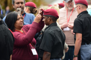 A woman adjusts the maroon beret of a young cadet in uniform during an outdoor gathering at Valley Forge Military College, surrounded by others, some also wearing berets and name badges.