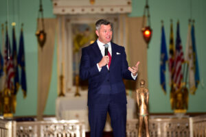 A man in a dark blue suit speaks into a microphone onstage, gesturing with one hand. Behind him are flags, lit candles, and ornate decorations. The event honors leadership at Valley Forge Military College; a gold statue stands before the stage.
