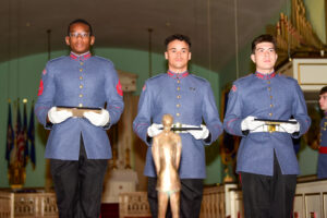 Three young cadets from Valley Forge Military College stand in a row, holding plaques and wearing white gloves inside a decorated hall of flags and an organ. A gold statue is blurred in the foreground, highlighting their commitment to leadership.