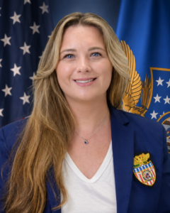 A woman with long blonde hair in a blue blazer with a badge, smiling, stands in front of U.S. flags with stars and an eagle emblem.