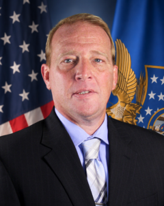 A man in a suit and tie poses in front of U.S. and blue government flags, with a serious expression.