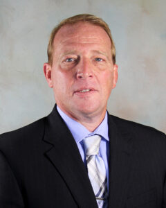 A man with short light brown hair, wearing a dark suit, light blue shirt, and patterned tie, poses in front of a neutral, light-colored background.