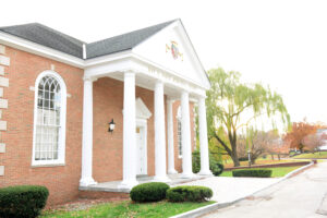 A brick building with tall white columns at the entrance, arched windows, and a triangular pediment stands amid autumn trees at Valley Forge Military College, reflecting its tradition of leadership and cadet excellence under a bright, clear sky.