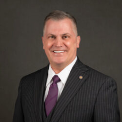 A man in a dark pinstripe suit, white shirt, and purple tie smiles at the camera against a plain dark background, reflecting the leadership and discipline instilled by his time as a cadet at Valley Forge Military College.