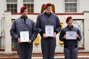 Three uniformed cadets wearing maroon berets stand outdoors, holding certificates and posing for a photo. They are in front of a white brick wall with gold and red accents, embodying the leadership values of Valley Forge Military College.