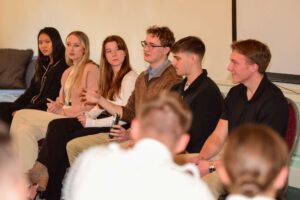 Six young adults, including a cadet from Valley Forge Military College, sit in a row as one gestures while speaking. The group appears engaged in a leadership panel discussion or meeting, with a few audience members visible in the foreground.