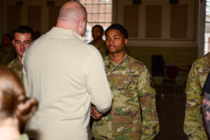 A person in a light sweater shakes hands with a Valley Forge Military College cadet in uniform at an indoor event, while other soldiers stand nearby, highlighting the spirit of leadership and camaraderie.