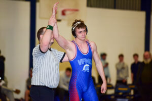 A referee raises the arm of a cadet wrestler in a blue and red singlet, signaling his victory. Showing calm leadership, he stands slightly tired amid people and blurred gym equipment, evoking the spirit of Valley Forge Military College.