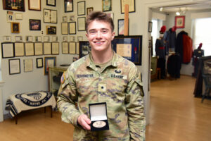 A smiling U.S. Army soldier in uniform stands indoors, displaying a medal in an open box. The background features framed photos and documents, with museum exhibits celebrating Leadership and Valley Forge Military College achievements.