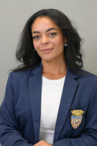 A woman with long dark hair, wearing a navy blazer with a crest on the pocket and a white shirt, stands in front of a plain gray background, smiling softly.
