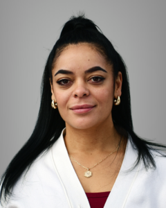 A woman with long black hair in a high ponytail, wearing gold hoop earrings, a gold necklace, a white top, and a red shirt underneath, poses against a plain gray background.