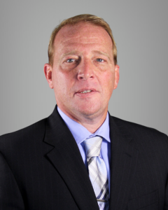 A middle-aged man with short blonde hair wearing a black suit, light blue dress shirt, and patterned tie, posing against a plain gray background.