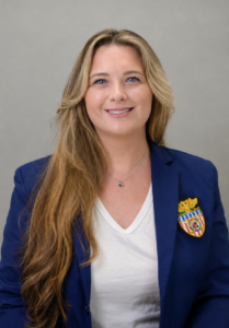 A woman with long blonde hair, wearing a blue blazer with a badge and a white shirt, smiles at the camera against a plain gray background.