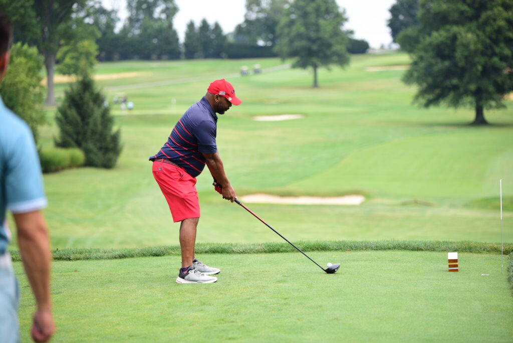 A man wearing a red cap, red shorts, and a navy striped shirt prepares to hit a golf ball on a green golf course; another person stands partially visible in the foreground.