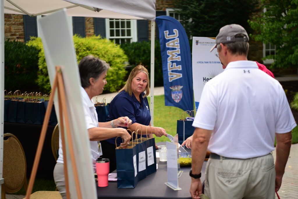 People stand at an outdoor registration table with gift bags, a sign, and a VFMAC banner. One woman hands a bag to a man, while another woman sits behind the table. Trees and a building are in the background.