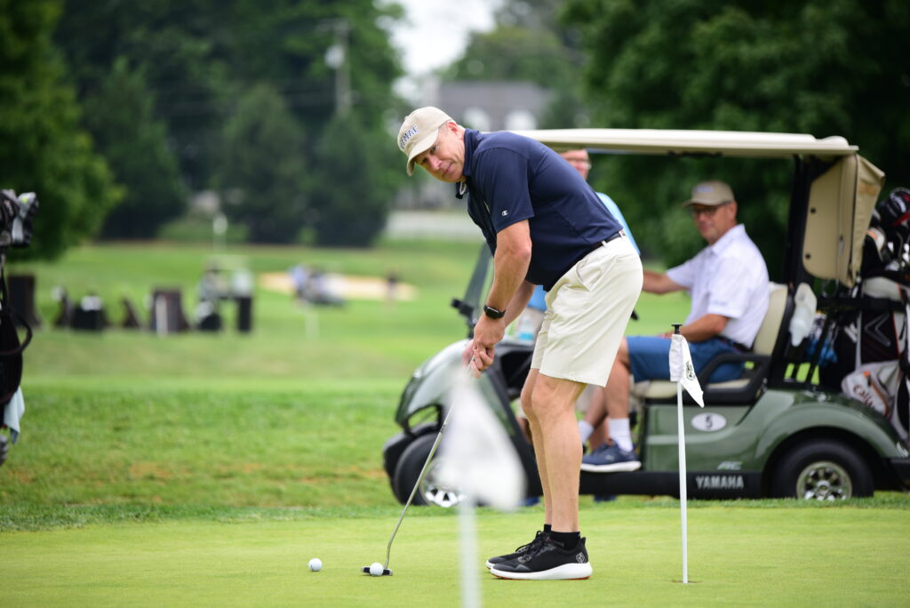 A man in a navy shirt and khaki shorts is putting on a golf green, with two men sitting in a golf cart in the background. Trees and golf equipment are visible around the course.