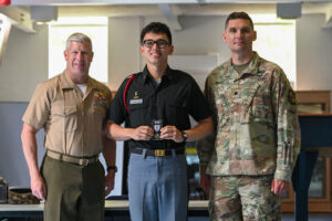Three men stand indoors, two in military uniforms and one in a black shirt and gray pants holding a medal. They are posing for a photo, smiling, with a blurred background.