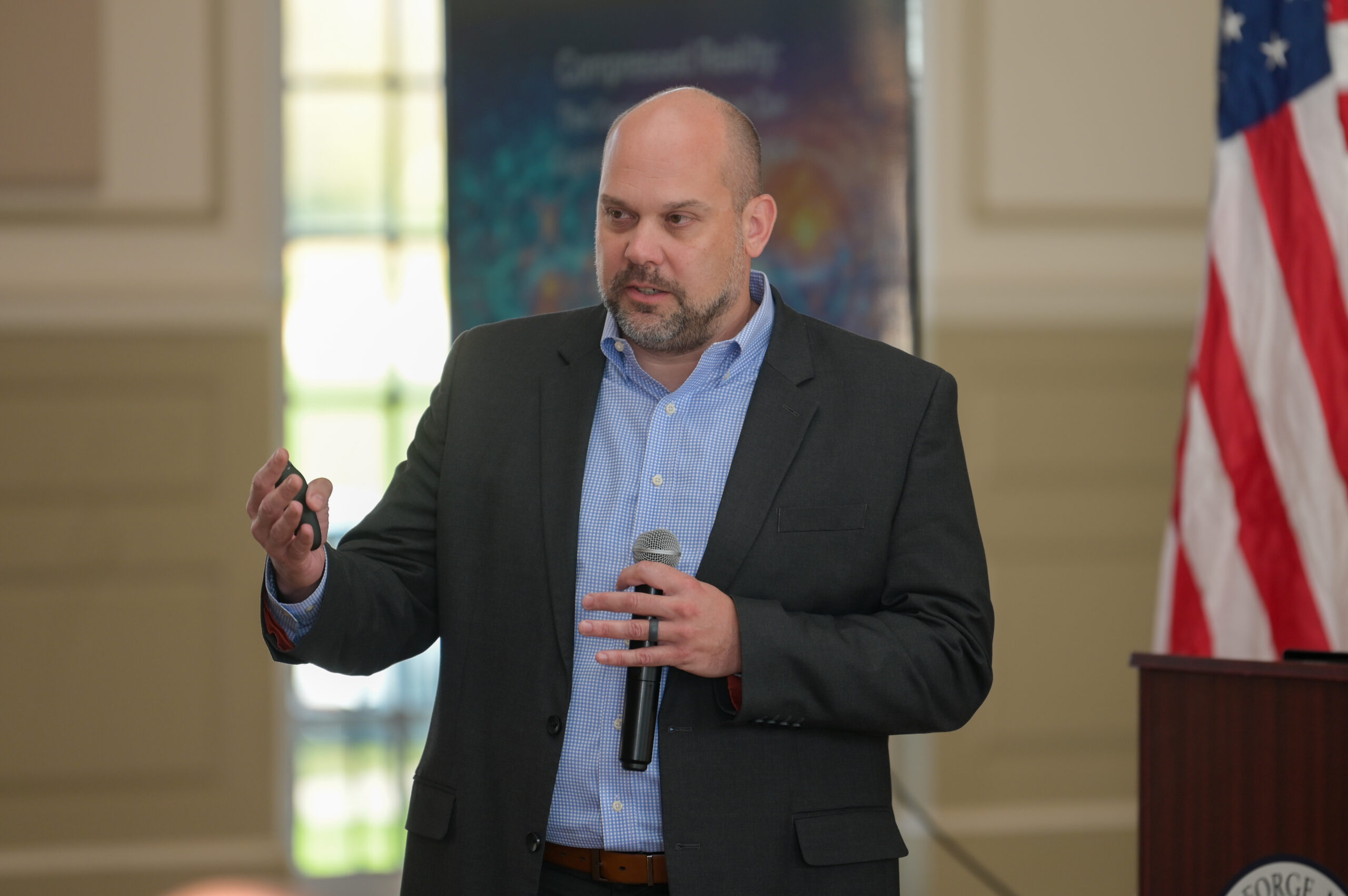 A man in a suit holding a microphone and gesturing while speaking at an indoor event, with an American flag and podium in the background.