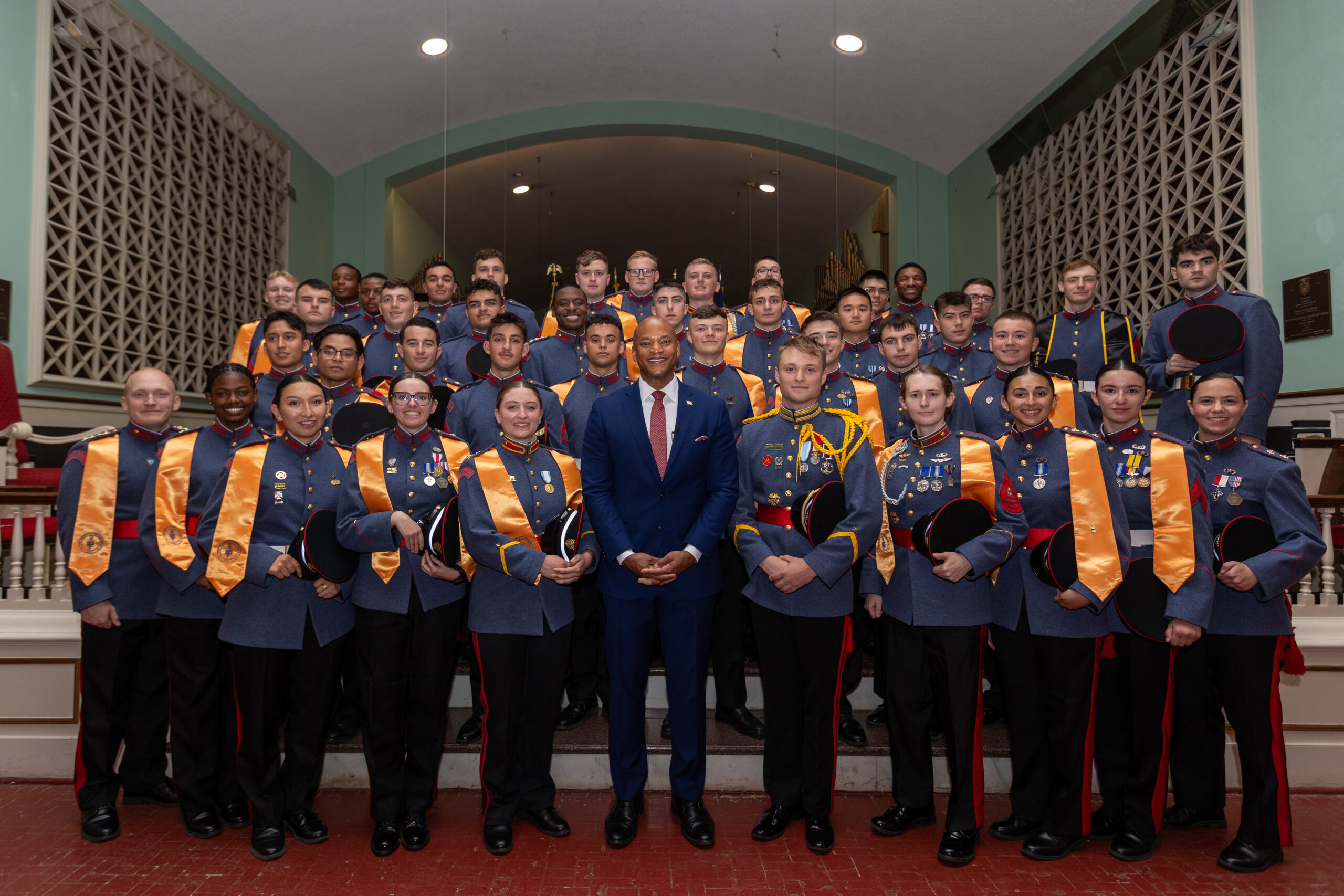 A large group of uniformed cadets with orange stoles pose on a staircase around a man in a blue suit, smiling for a formal group photo inside a building with decorative walls.