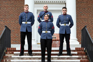 Four uniformed cadets stand on brick steps in front of a building with white columns. Three men and one woman stand in formation, facing forward with their hands behind their backs, wearing blue jackets and dark pants.