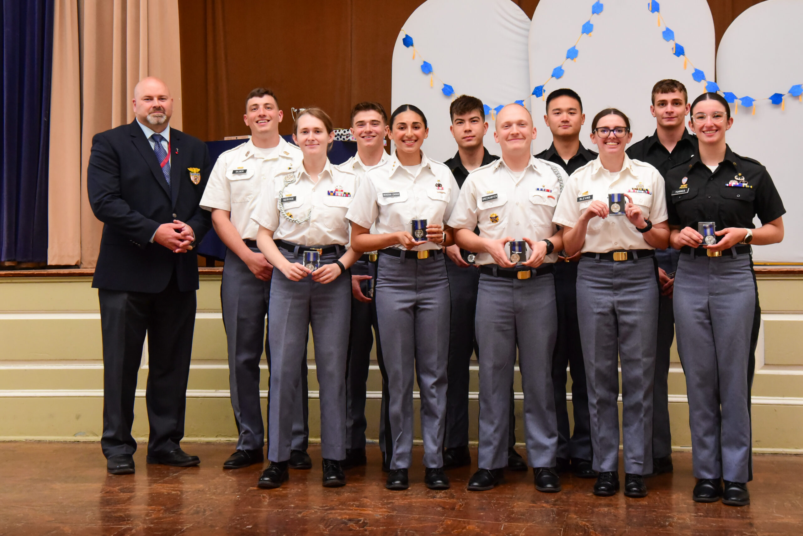 A group of cadets in uniform stand in a line indoors, holding medals, with an adult in a suit beside them. They are posing for a photo on a stage with decorations in the background.