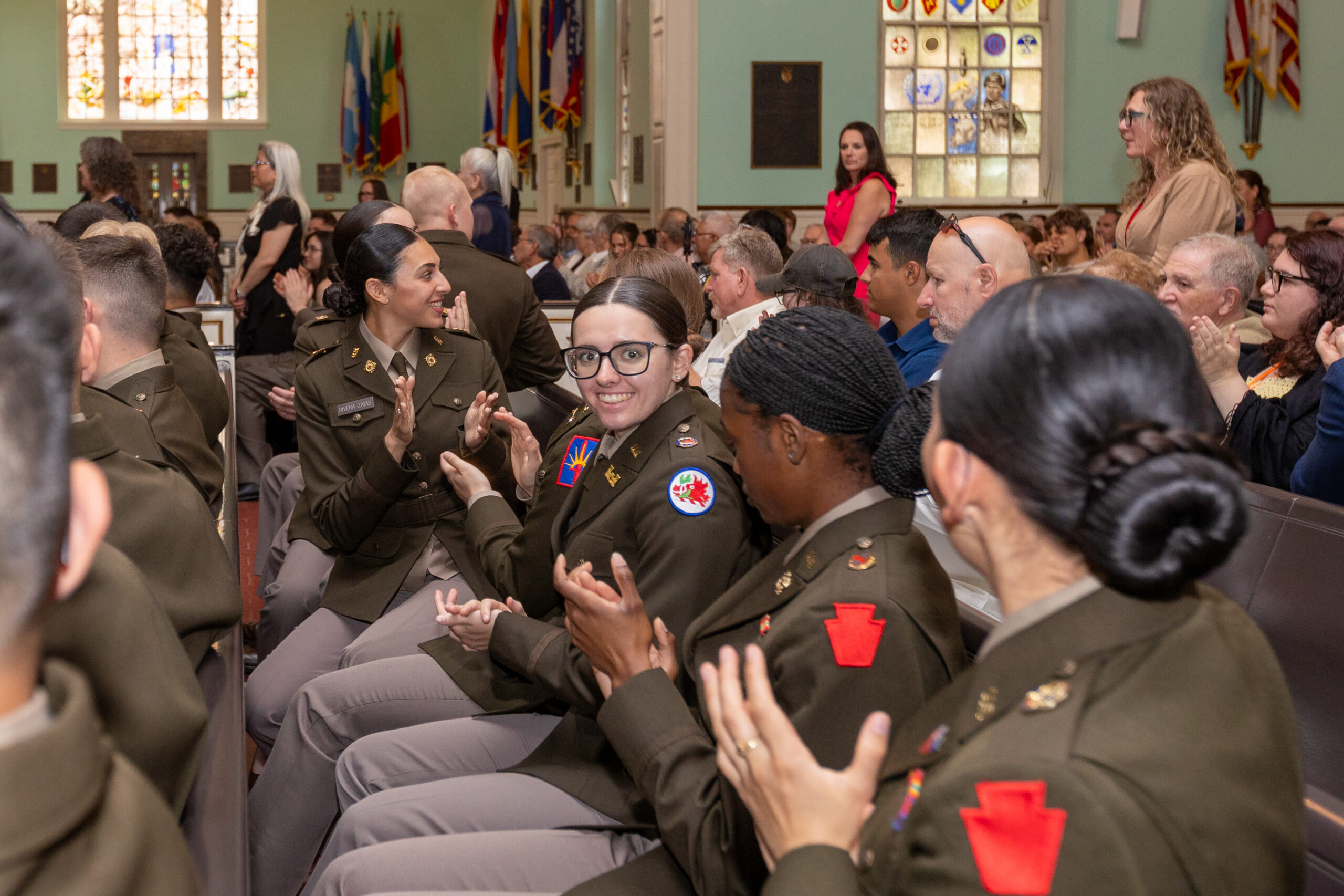 A group of uniformed military personnel sit and clap in an indoor auditorium filled with people, some of whom are also applauding. Flags and stained glass windows are visible in the background.
