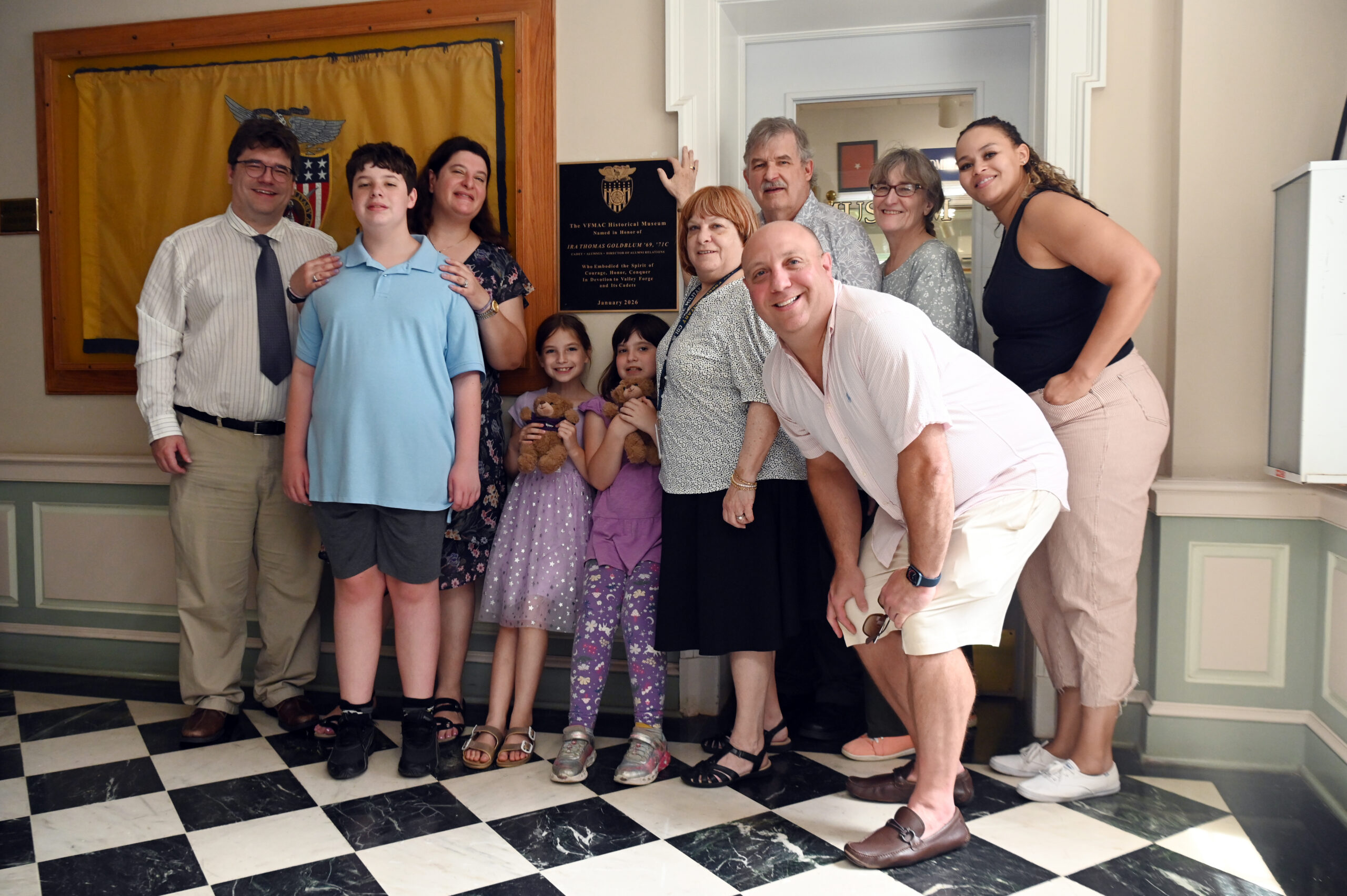 A group of ten people, including adults and children, pose and smile together indoors on a black-and-white checkered floor in front of a wall with plaques and a yellow display case.