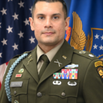 A soldier in formal military uniform stands in front of American and blue flags. He wears medals, badges, and a name tag reading BEDRIN. He has short dark hair and a serious expression.