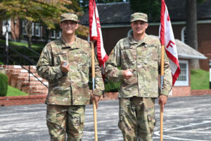 Two soldiers in camouflage uniforms hold red and white flags and baseballs while standing outdoors on a paved area, smiling at the camera with buildings and greenery in the background.