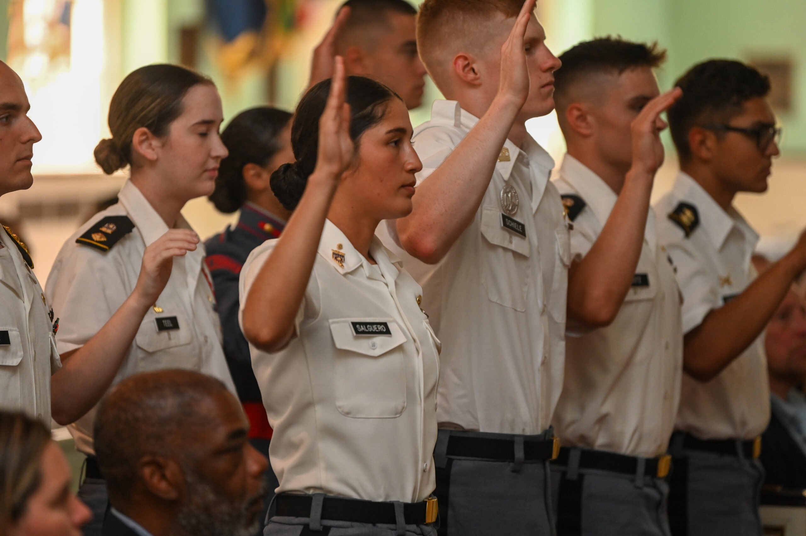 A group of cadets in uniform stand with their right hands raised, taking an oath during a formal ceremony indoors.