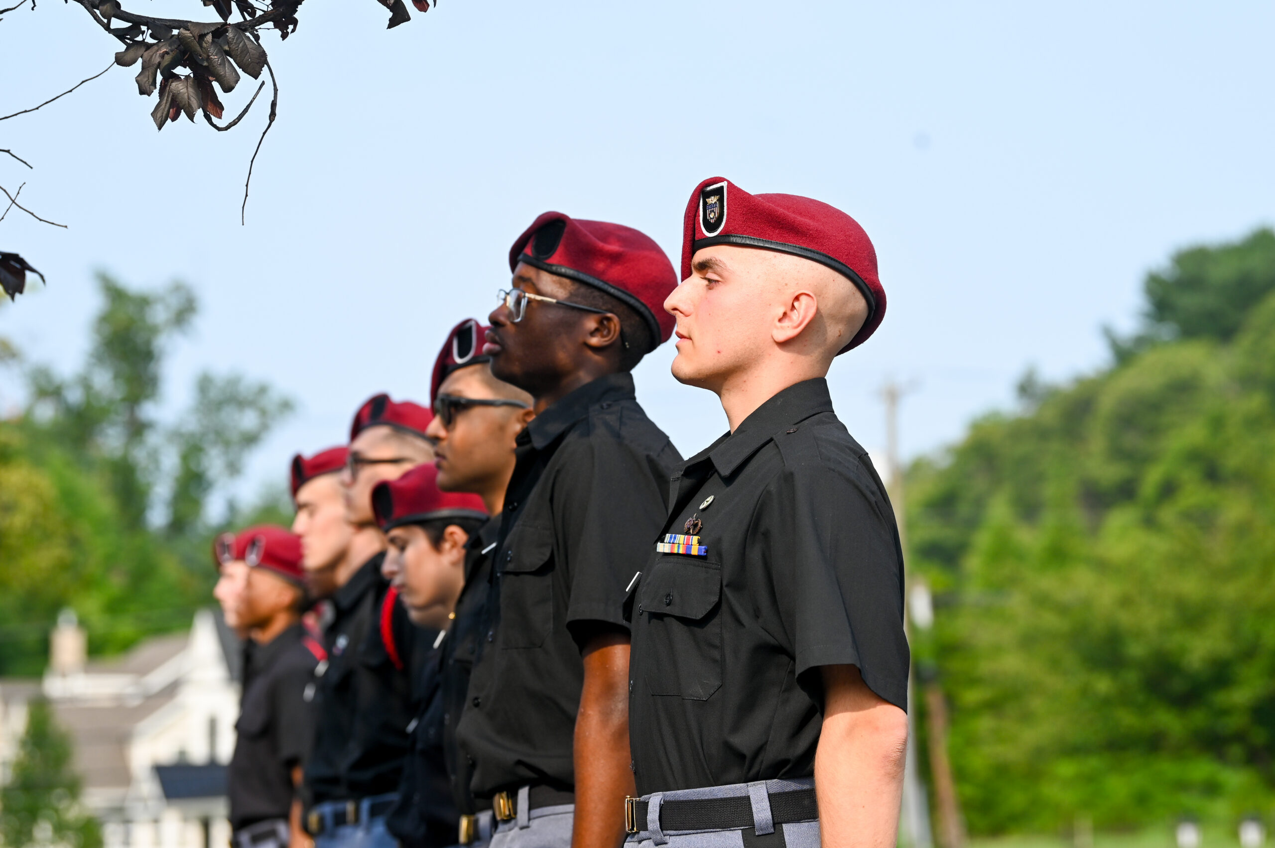 A line of uniformed cadets wearing maroon berets stand outdoors at attention, looking forward. Trees and buildings are visible in the background under a clear sky.