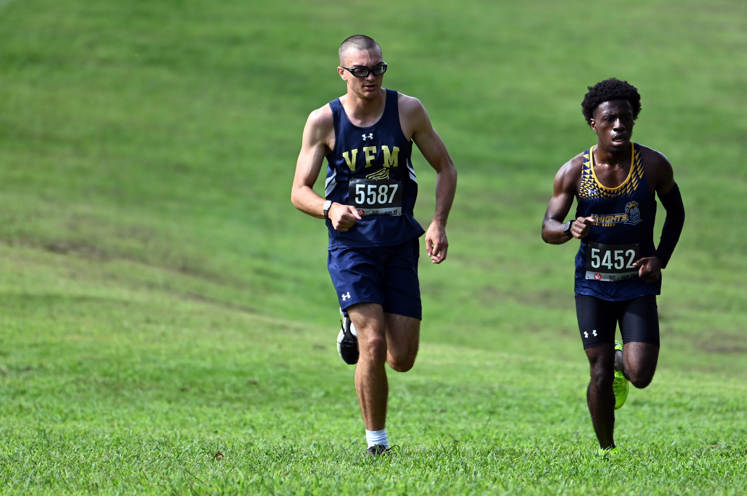 Two male runners wearing race bibs run uphill on grass during a race. The runner on the left wears sunglasses and a navy uniform, while the runner on the right wears a black and yellow uniform.