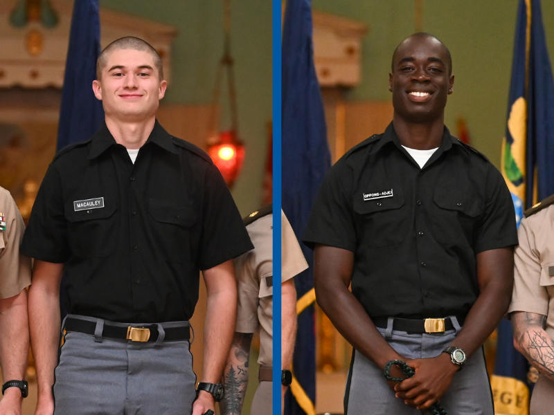 Two men in military-style uniforms standing side-by-side in front of flags.